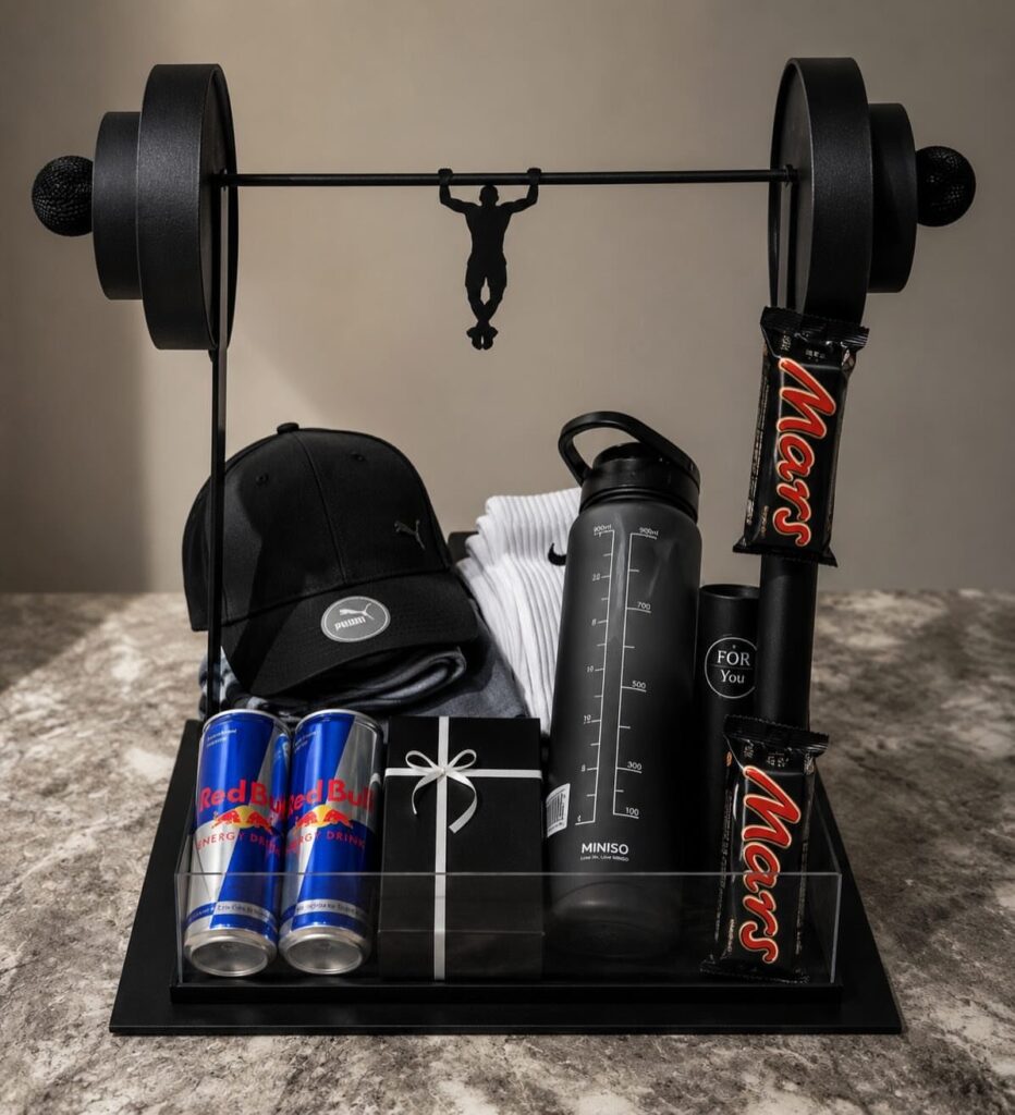 Fitness gift set with barbell frame, water bottle, Red Bull, and chocolate bars.