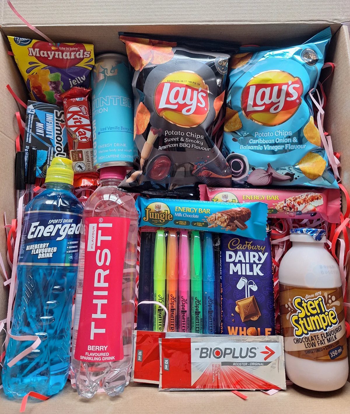Cardboard box filled with snacks, drinks, and markers on pink shredded paper.