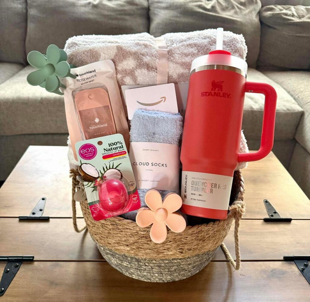 Woven gift basket containing a red tumbler, plush blanket, and beauty products on table