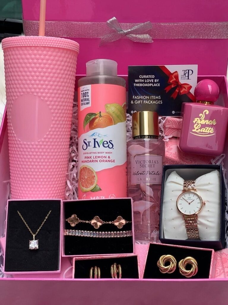 Pink gift box containing a tumbler, skincare, perfumes, and gold jewelry on pink filler.