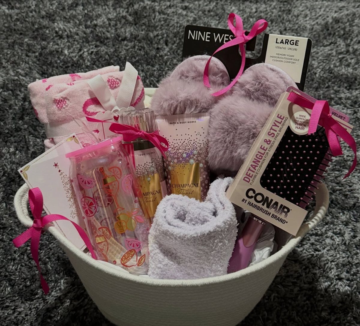 White basket filled with pink slippers, towels, a hairbrush, and champagne on grey rug.